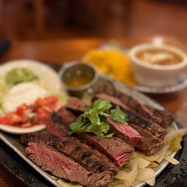 Sliced medium-rare steak garnished with cilantro, served over sautéed onions with small bowls of sauce and vegetables.