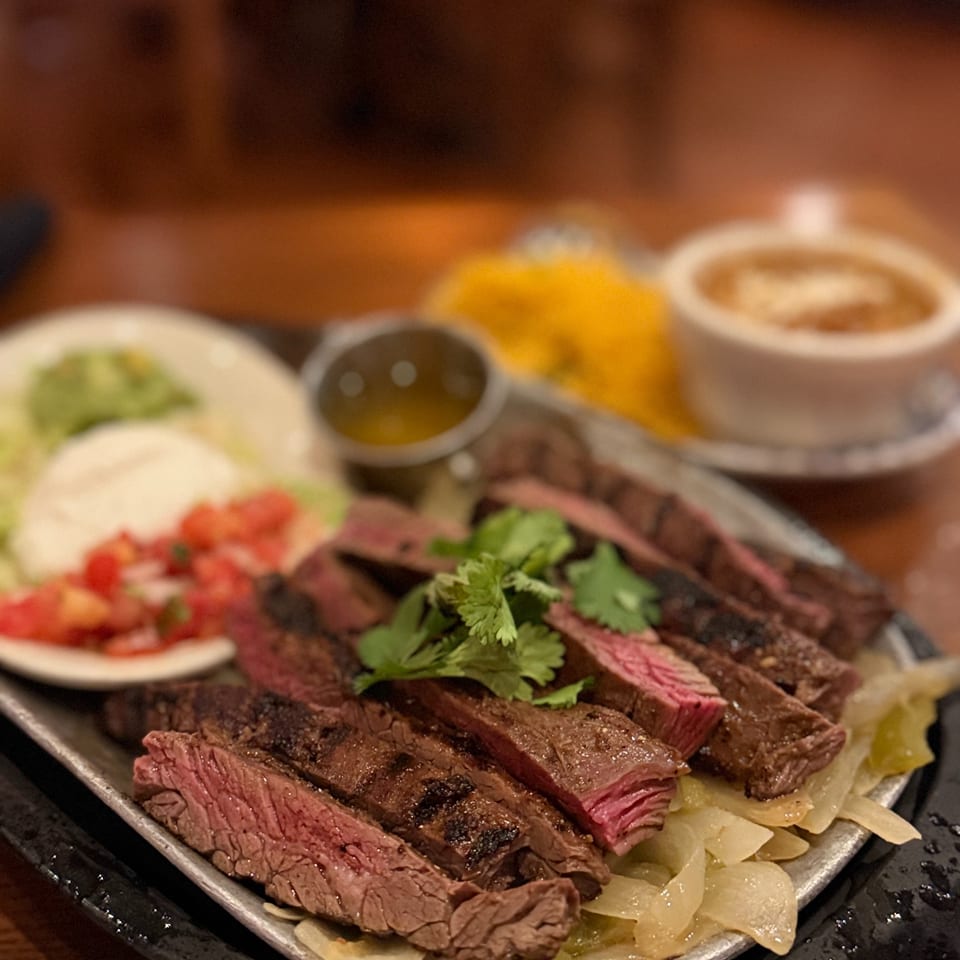 Sliced medium-rare steak garnished with cilantro, served over sautéed onions with small bowls of sauce and vegetables.