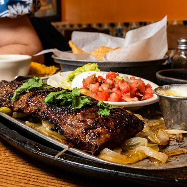 Sizzling fajita steak with onions, cilantro, pico de gallo, and guacamole on a wooden table.