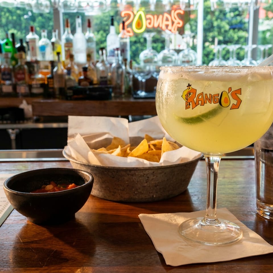A margarita, chips and salsa, and a menu on a wooden bar with a neon sign.